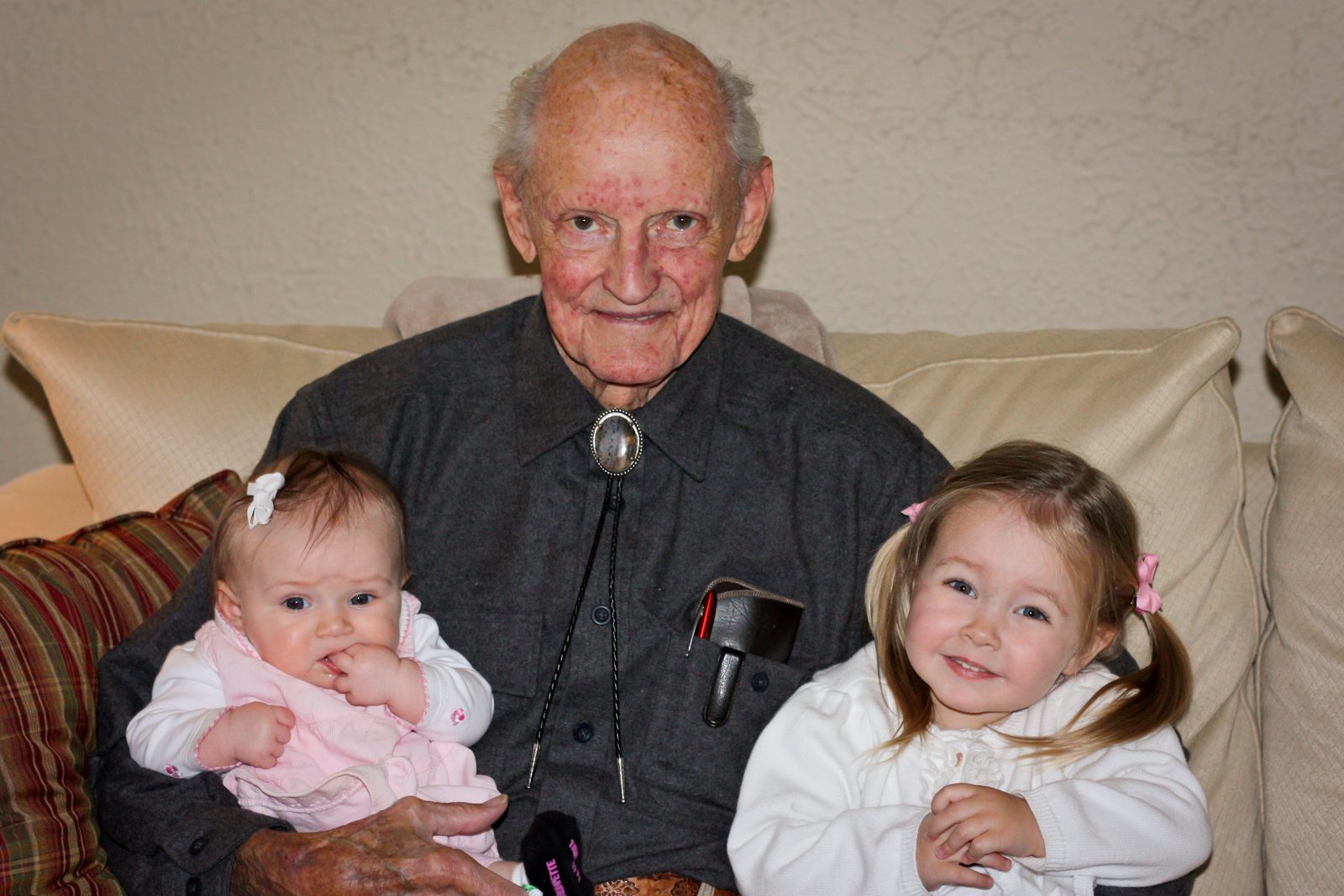 Duane in a bolo tie holding two of his great-grandchildren on the couch